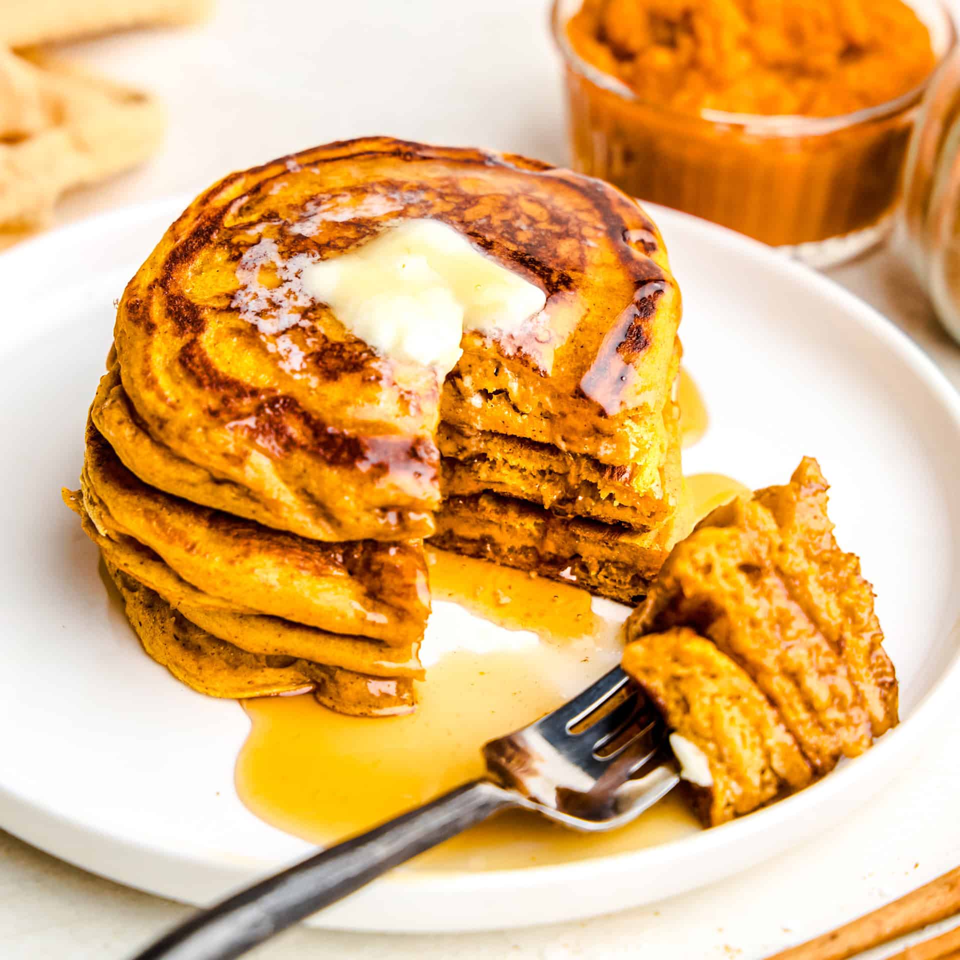 A stack of Pumpkin Pancakes on a plate with a slice taken out of them and cinnamon and pumpkin puree in the background.
