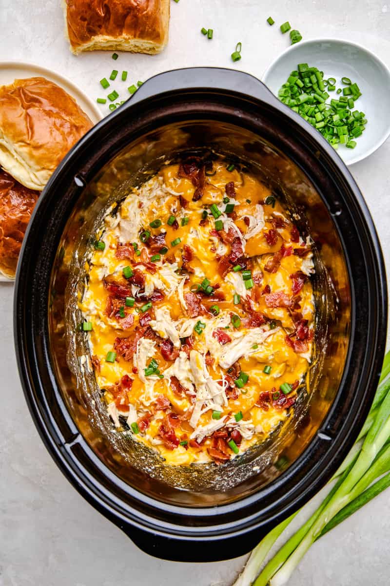 Overhead image of cheesy Slow Cooker Crack Chicken in a crockpot with rolls and green onions scattered.
