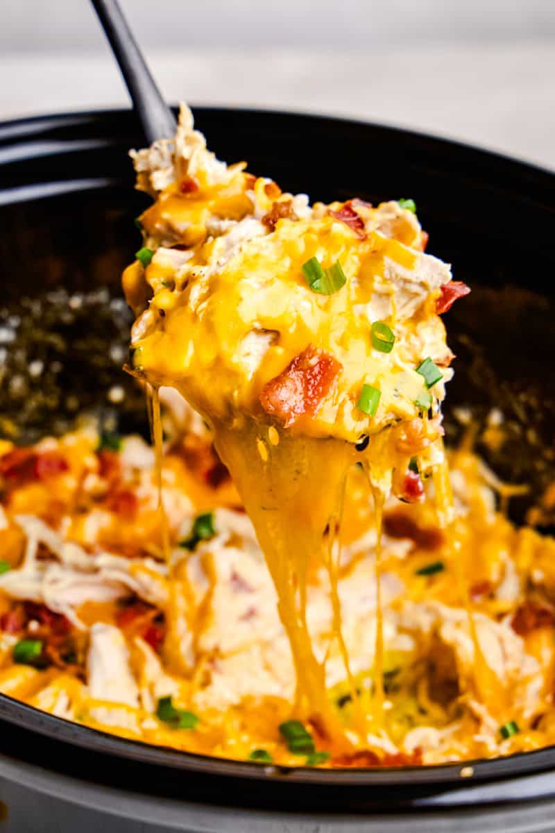 A serving spoon of cheesy Slow Cooker Crack Chicken being lifted out of a crockpot with a melty cheese pull.