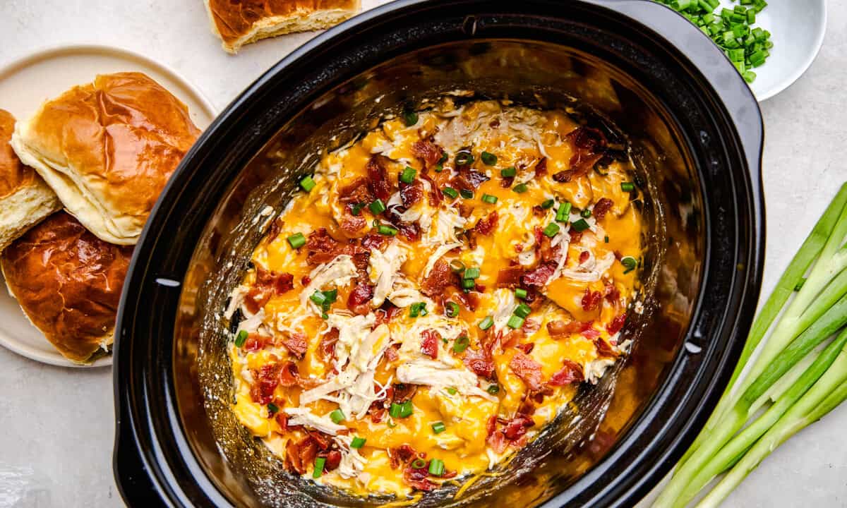 Overhead image of cheesy Slow Cooker Crack Chicken in a crockpot with rolls and green onions scattered.