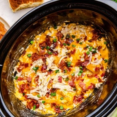 Overhead image of cheesy Slow Cooker Crack Chicken in a crockpot with rolls and green onions scattered.