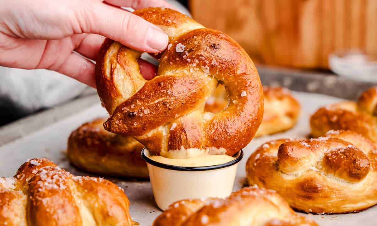 A hand holding a Soft Pretzel and dipping it into cheese sauce on a pan of freshly baked pretzels.