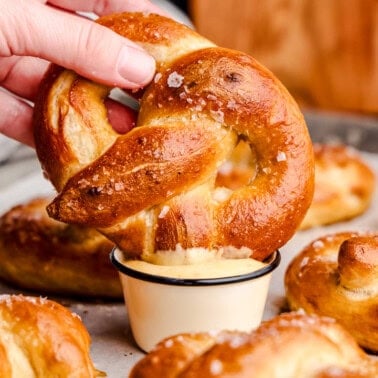 A hand holding a Soft Pretzel and dipping it into cheese sauce on a pan of freshly baked pretzels.