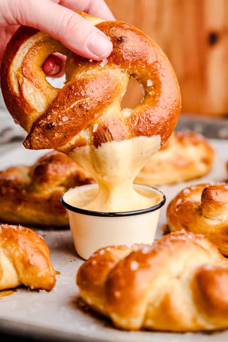 A hand holding a Soft Pretzel and dipping it into cheese sauce on a pan of freshly baked pretzels