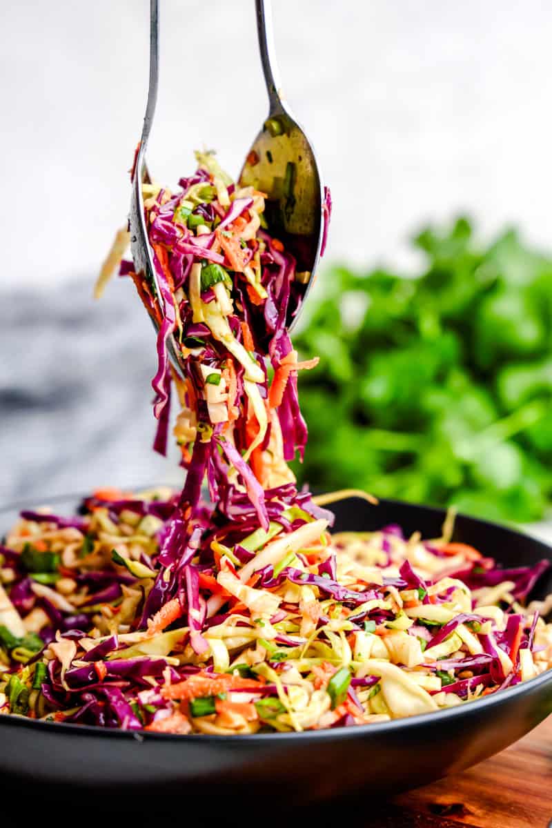 Two spoons lifting up and serving Spicy Sesame Coleslaw from a black serving bowl.