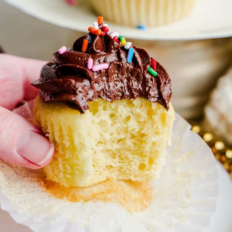 A hand holding a vanilla cupcake with chocolate frosting with a bite taken out of it.
