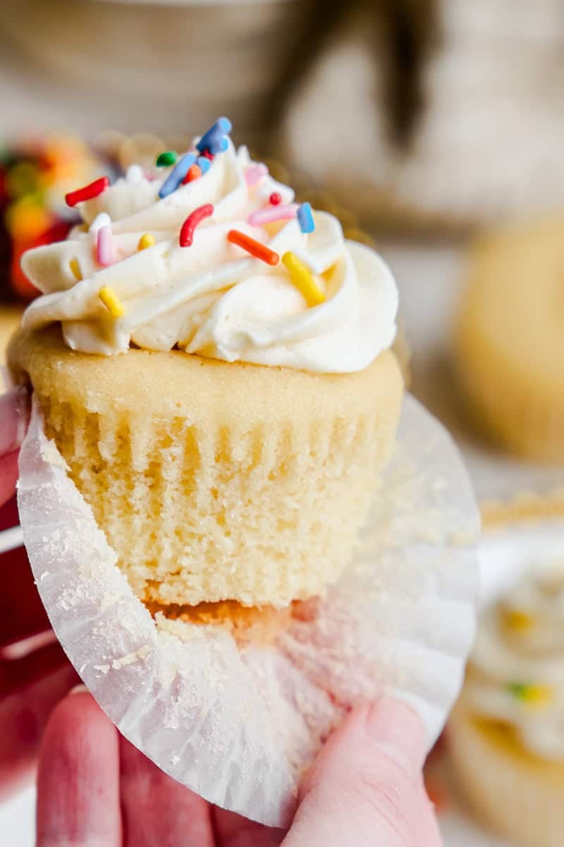 A hand opening the wrapper of a vanilla cupcake with white frosting and colorful sprinkles.