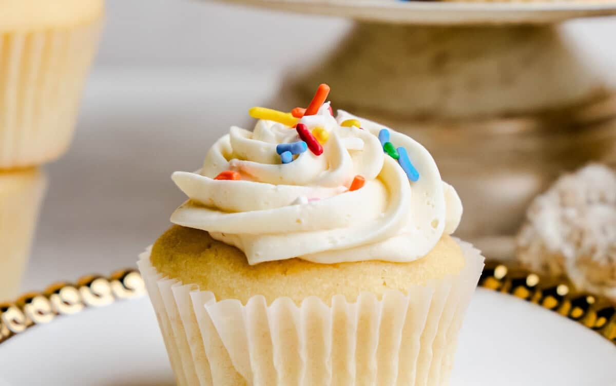 A vanilla cupcake on a plate with colorful sprinkles on the frosting.