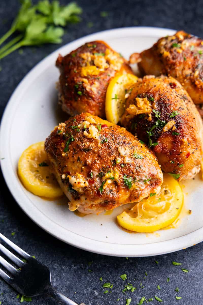 Four chicken thighs with lemon slices and garlic on a white plate.