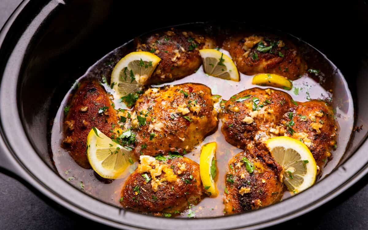 An overhead view into a slow cooker where there are chicken thighs that have been slow cooked in a buttery lemon garlic sauce and garnished with lemon slices.