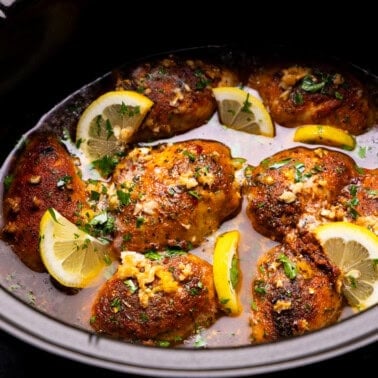An overhead view into a slow cooker where there are chicken thighs that have been slow cooked in a buttery lemon garlic sauce and garnished with lemon slices.