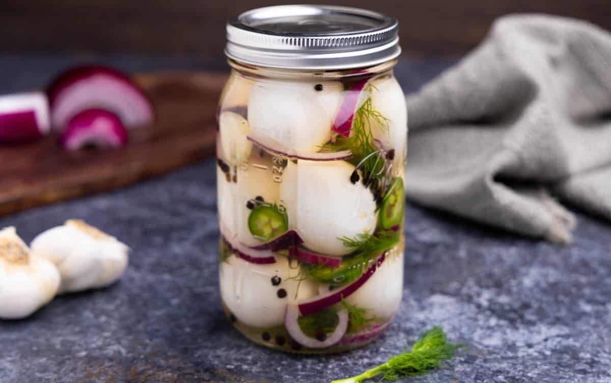 A jar of pickled eggs on a surface with red onion, black peppercorns, fresh dill, and jalapeno.