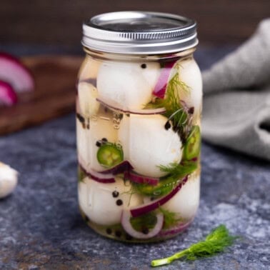 A jar of pickled eggs on a surface with red onion, black peppercorns, fresh dill, and jalapeno.