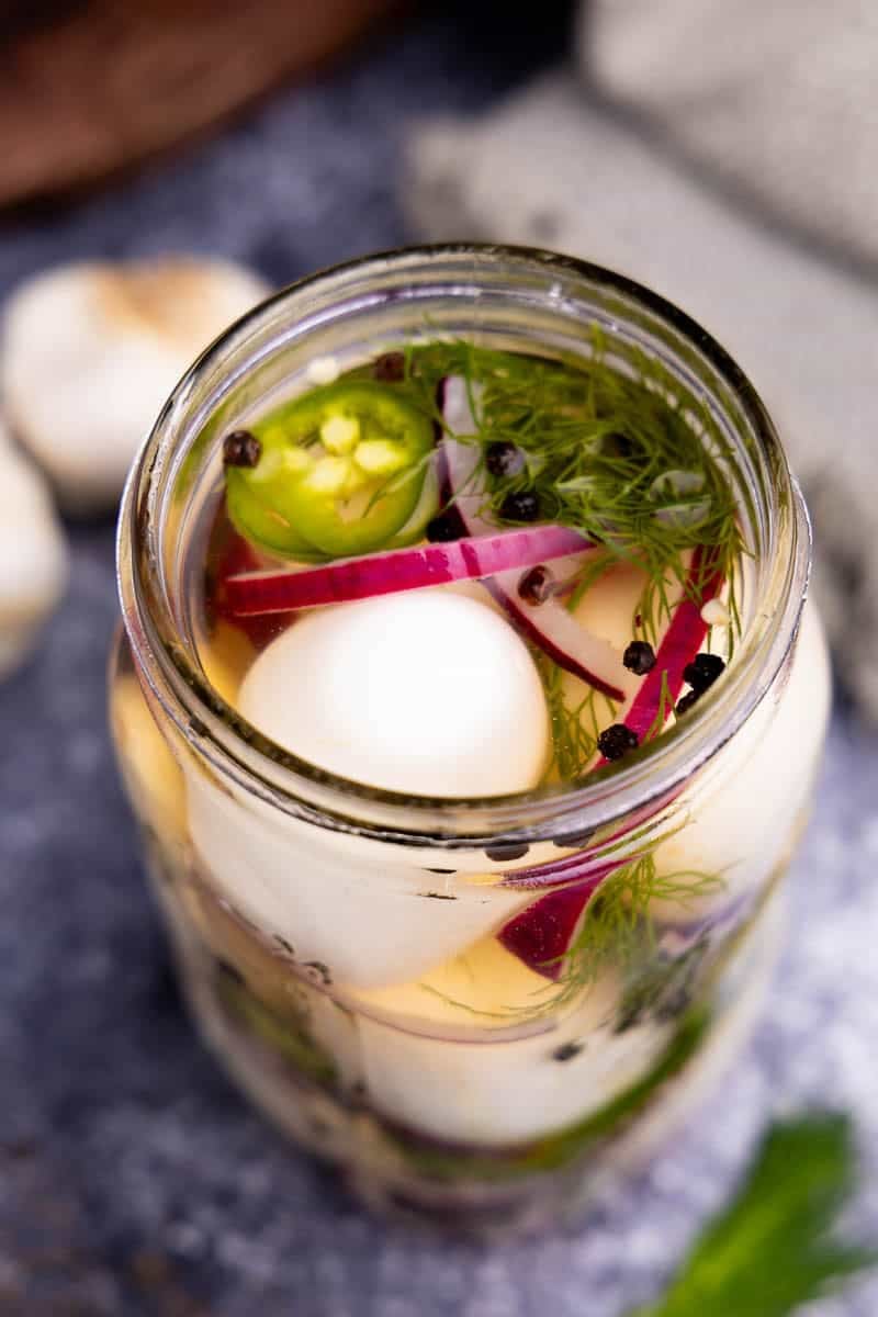 A peek into a jar of pickled eggs with red onion, black peppercorns, fresh dill, and jalapeno.