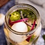 A close up peek into a jar of pickled eggs with red onion, black peppercorns, fresh dill, and jalapeno.