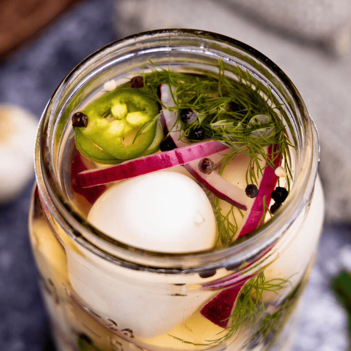 A close up peek into a jar of pickled eggs with red onion, black peppercorns, fresh dill, and jalapeno.