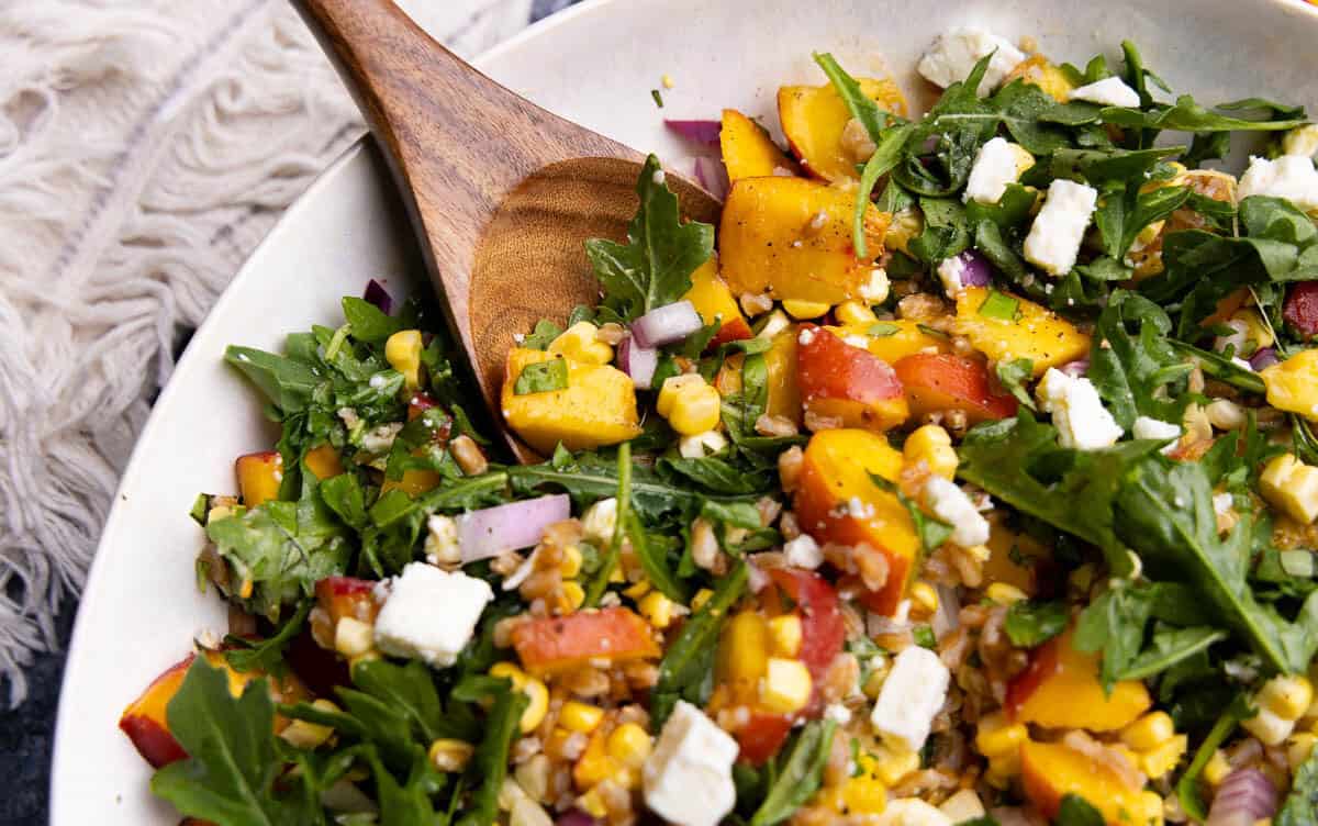 A close up view of a bowl of peach farro salad with arugula, feta cheese, red onion, and fresh corn. There's a wooden spoon resting in the bowl.