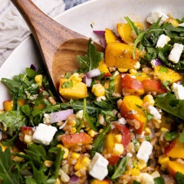 A close up view of a bowl of peach farro salad with arugula, feta cheese, red onion, and fresh corn. There's a wooden spoon resting in the bowl.