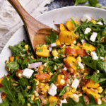 A close up view of a bowl of peach farro salad with arugula, feta cheese, red onion, and fresh corn.