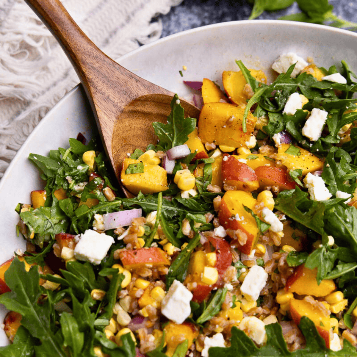 A close up view of a bowl of peach farro salad with arugula, feta cheese, red onion, and fresh corn.