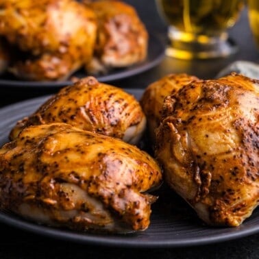 A pile of apple cider glazed chicken thighs on a black plate. There's an additional plate of chicken thighs in the background and mugs of apple cider.