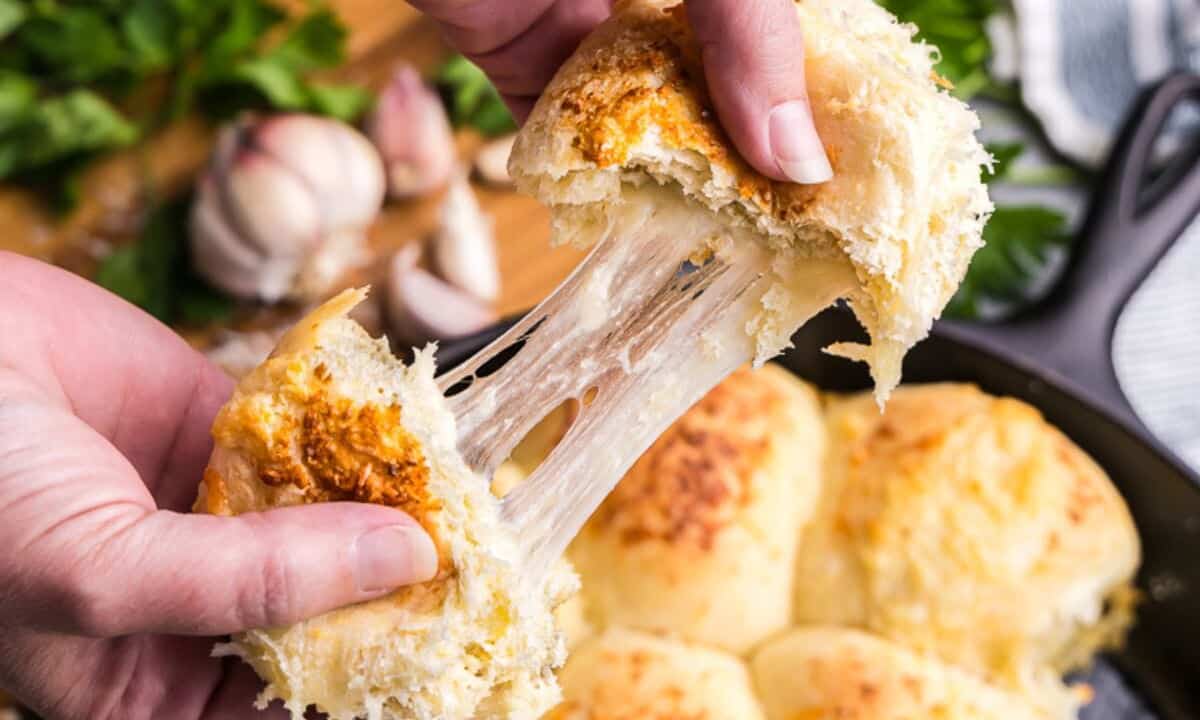 Hands pulling apart a cheese stuffed garlic butter dinner rolls, with a cast iron pan with the rest of the rolls behind. Gooey cheese shows being stretched apart.