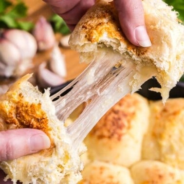 Hands pulling apart a cheese stuffed garlic butter dinner rolls, with a cast iron pan with the rest of the rolls behind. Gooey cheese shows being stretched apart.