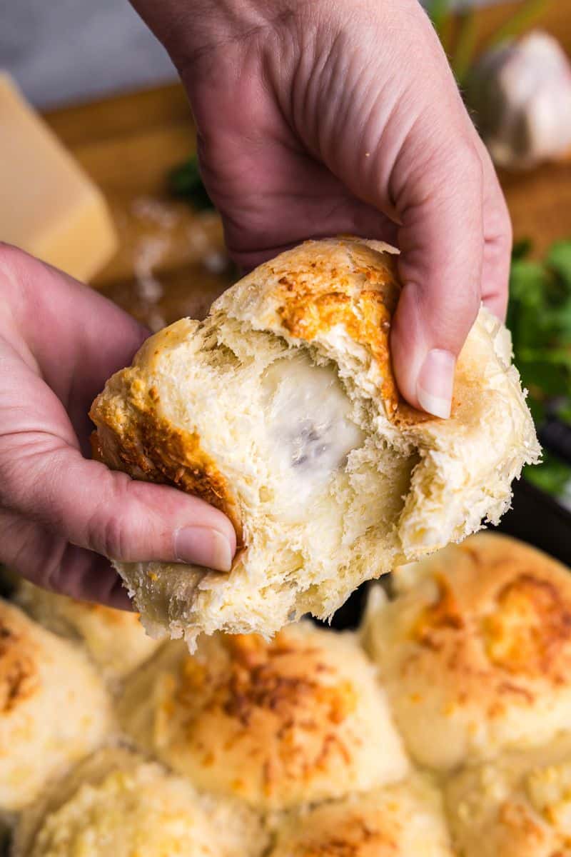 Hands pulling apart a cheese stuffed garlic butter dinner rolls, with a cast iron pan with the rest of the rolls behind. The cheese is just starting to ooze out.