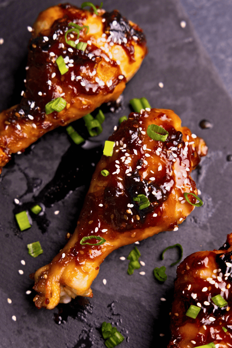 A closeup view of a chicken drumstick on a black platter that have been glazed in Korean BBQ sauce and garnished with sliced green onion and sesame seeds.