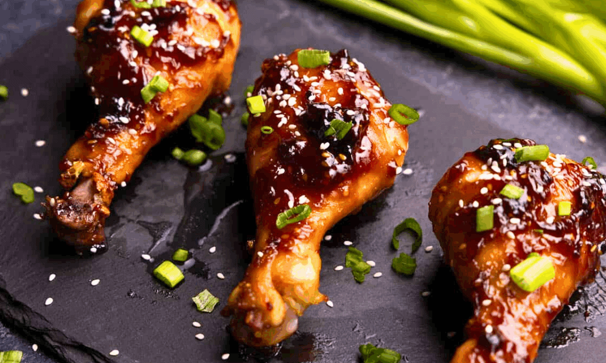 A closeup view of 3 drumsticks on a black platter that have been glazed in Korean BBQ sauce and garnished with sliced green onion and sesame seeds.