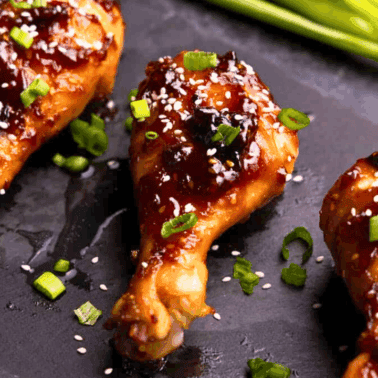 A closeup view of 3 drumsticks on a black platter that have been glazed in Korean BBQ sauce and garnished with sliced green onion and sesame seeds.