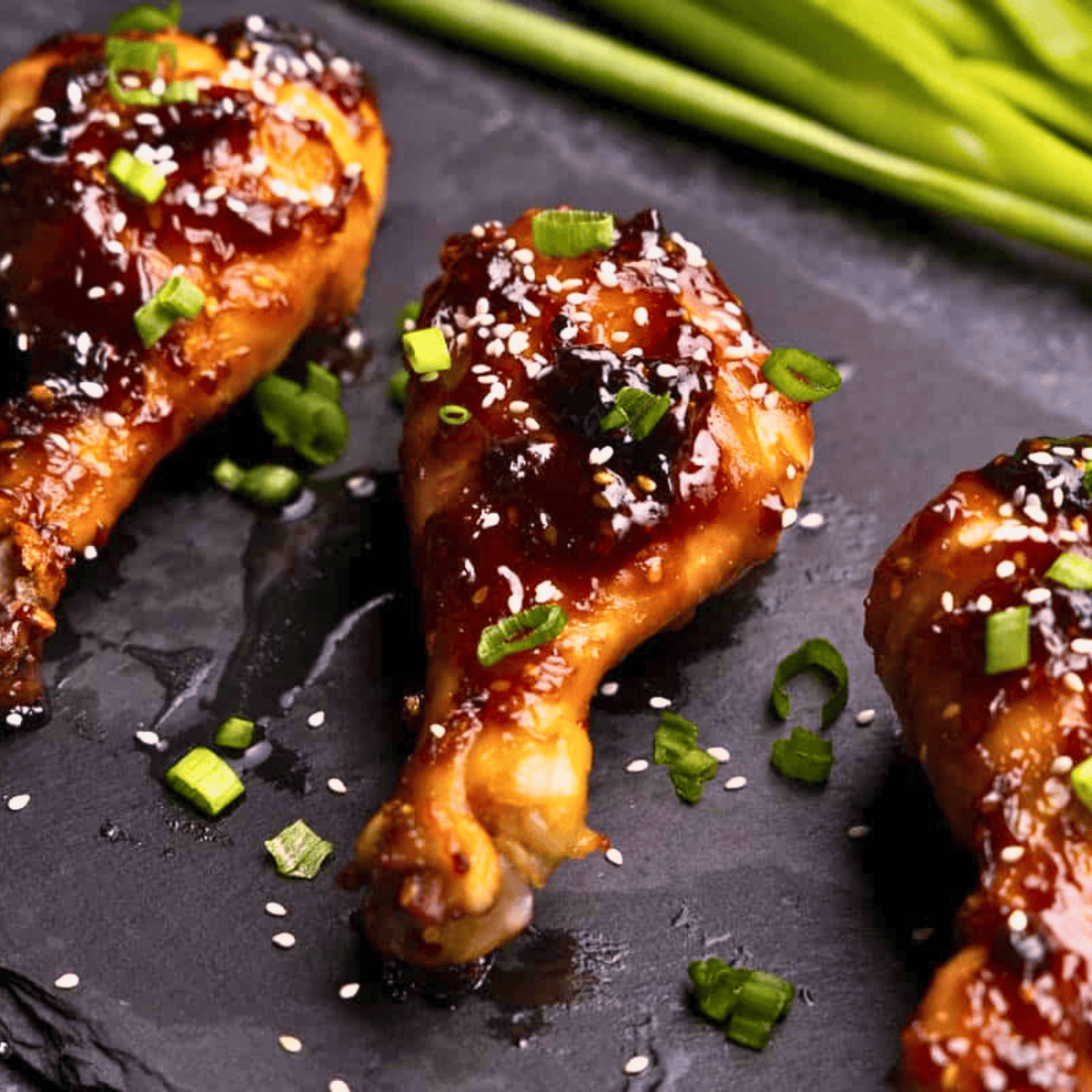 A closeup view of 3 drumsticks on a black platter that have been glazed in Korean BBQ sauce and garnished with sliced green onion and sesame seeds.