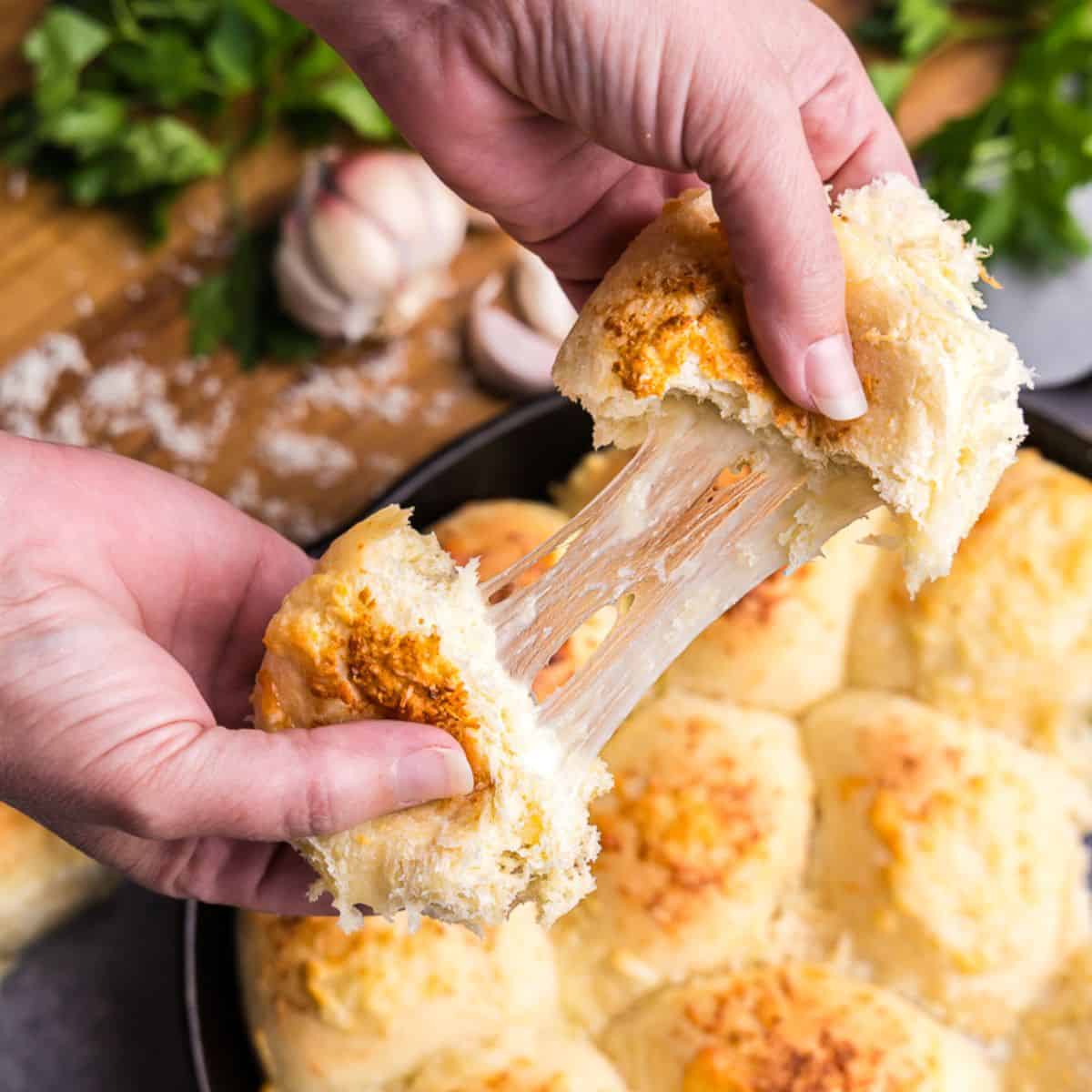 Hands pulling apart a cheese stuffed garlic butter dinner rolls, with a cast iron pan with the rest of the rolls behind. Strings of cheese are being stretched as the roll is pulled apart.