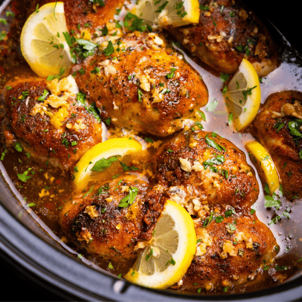 A closeup view of lemon garlic butter chicken thighs in the crock of a slow cooker, garnished with slices of lemon.