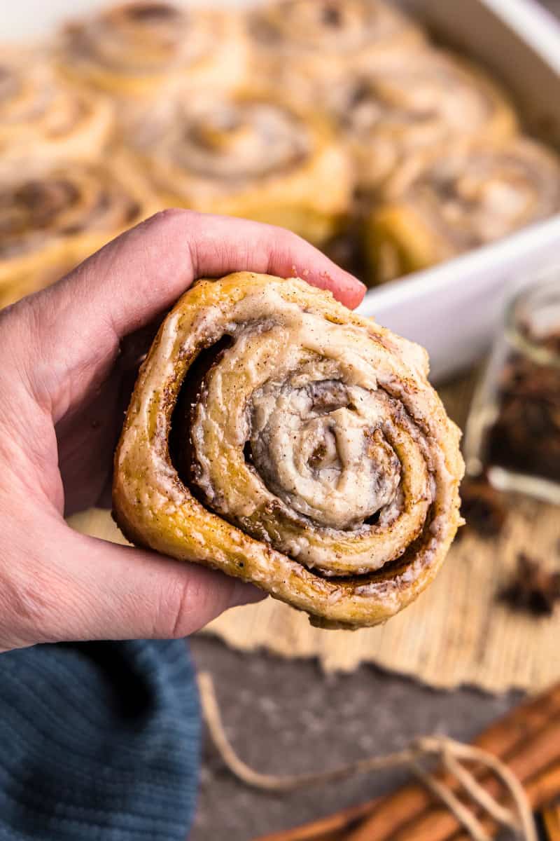 A hand holding a chai latte cinnamon roll. The pan of sweet rolls is out of focus in the background.