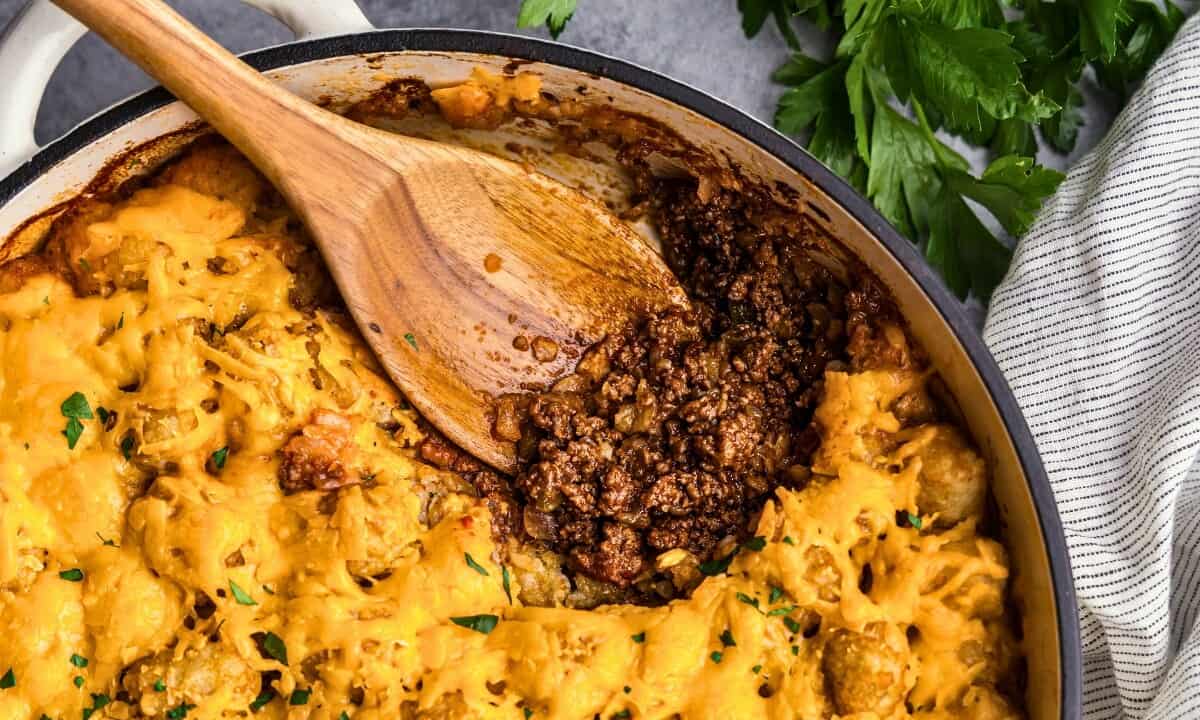 An overhead view of a sloppy joe tater tot casserole. The first helping has been scooped from the pan revealing the saucy sloppy joe ground beef beneath layers of crispy tater tots. A wooden serving spoon is resting in the pan.
