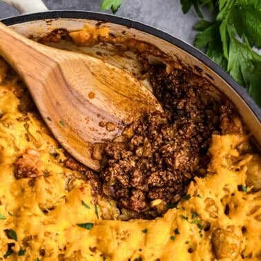 An overhead view of a sloppy joe tater tot casserole. The first helping has been scooped from the pan revealing the saucy sloppy joe ground beef beneath layers of crispy tater tots. A wooden serving spoon is resting in the pan.